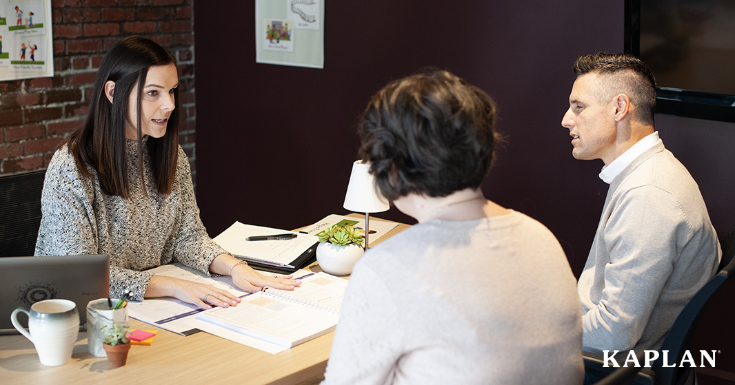 Woman seated at desk conducting a meeting with a man and a woman. 