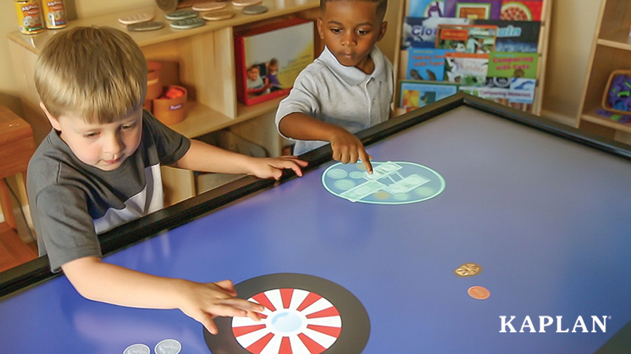 Two young boys play an educational game as they tap on the screen of a large, horizontal, interactive, tv panel.