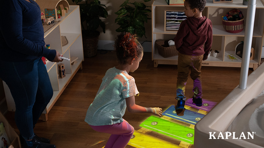 A young boy and a young girl take turns hopping across a colorful xylophone projected onto the wood floor, while a female teacher controls the projector with a remote control.