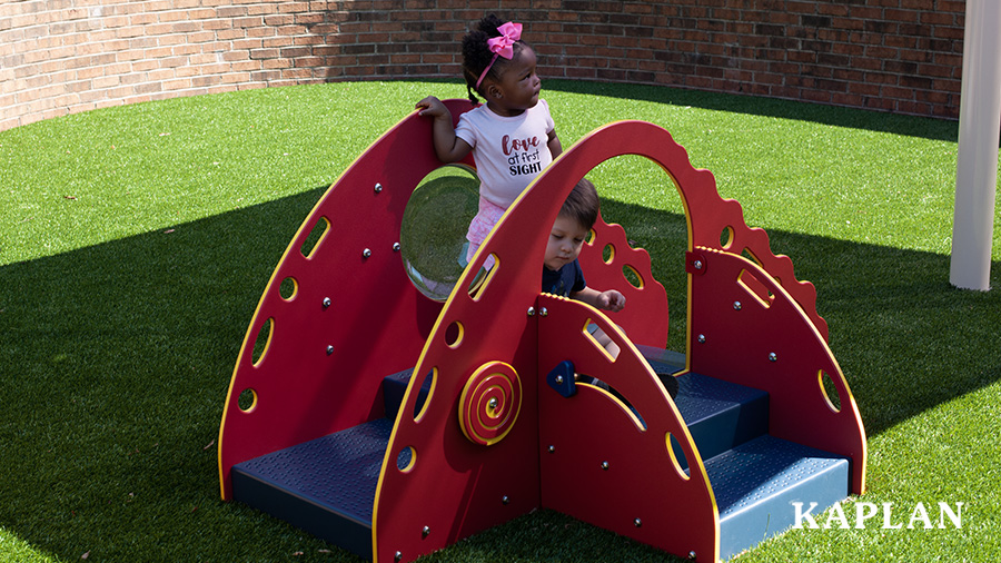 A young toddler girl and boy climb on a red and blue climber, with low steps, under a playground shade. 