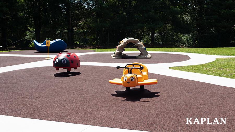 An image of a playground with rubber mulch surfacing, a yellow bumblebee bouncer, a red ladybug bouncer, a gray climbing wall, and a blue inclusive spinner.