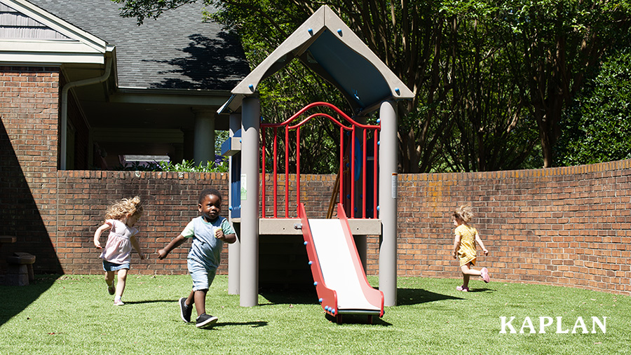 Three preschool age children run around a grey and red slide on a grass covered playground.