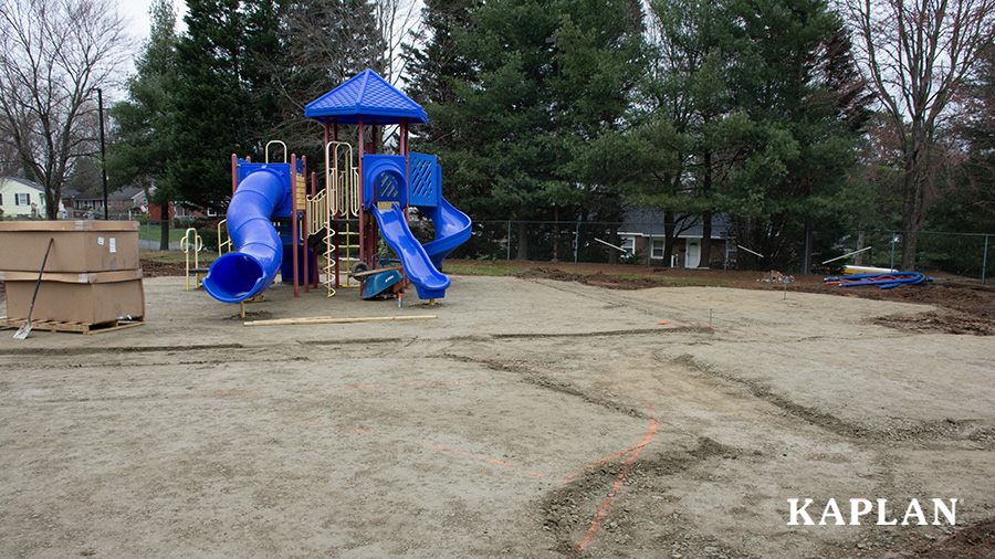 A playground site with boxes, construction materials, and a blue, red, and yellow playground climber in a dirt cover space, grated and leveled for surfacing installation.
