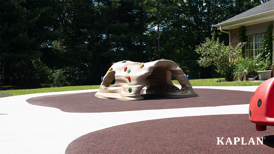 A plastic rock climbing structure surrounded by smooth surfacing, sidewalks, grass, trees, and landscaping..