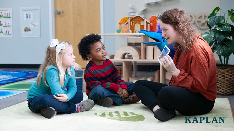 An early childhood teacher sits on a carpeted area of her classroom, holding up a card. Two children sit in front of the teacher, both looking at the card in her hand. 