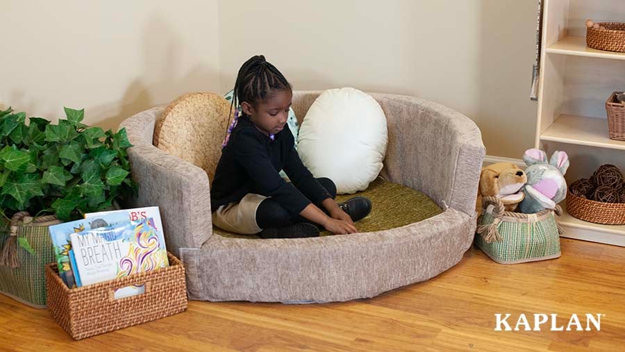 A young girl wearing a black shirt sits in the Kaplan Cozy Nest couch which is positioned in the corner of an early childhood classroom. 