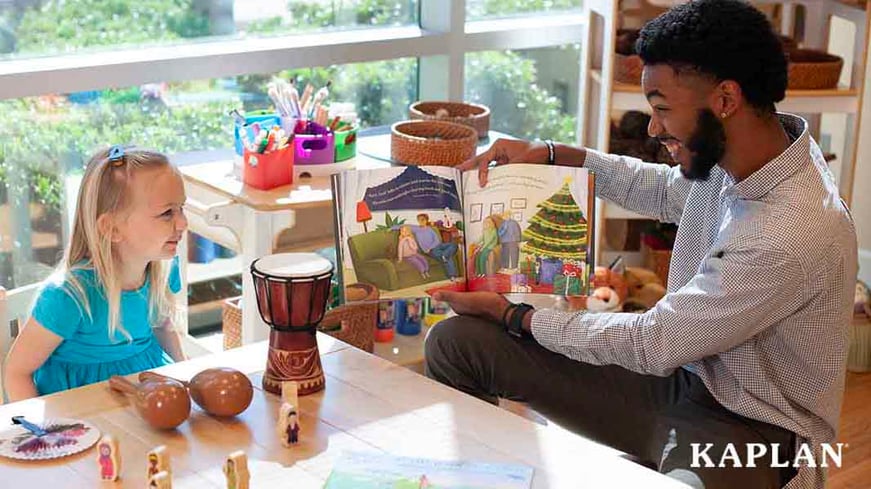 A male early childhood teacher sits at a table, holding a book, while a young girl in a blue dress looks at the book. 