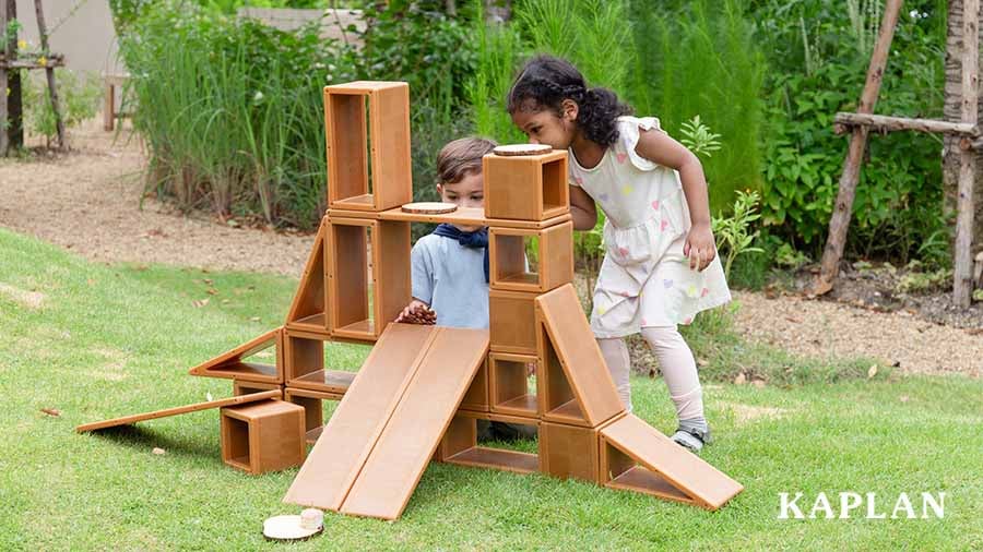 Two children build a tall structure on a grassy outdoor area using the Kaplan All-Weather Blocks. The children are pushing natural elements, like pinecones, down a ramp that is situated in the middle of the wooden block structure. 