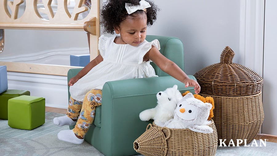 A preschool-aged girl sits in a green vinyl chair and reaches to grab a stuffed animal from a wicker basket beside the chair