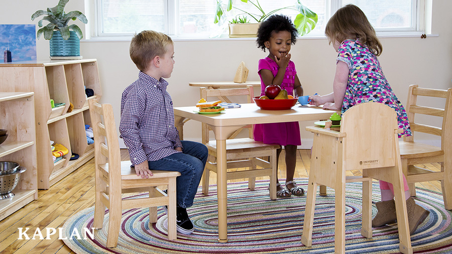 Three preschool-aged children (two girls and one boy) eat a pretend meal at a light brown, plastic table surrounded by three wooden classroom chairs and one wooden high chair