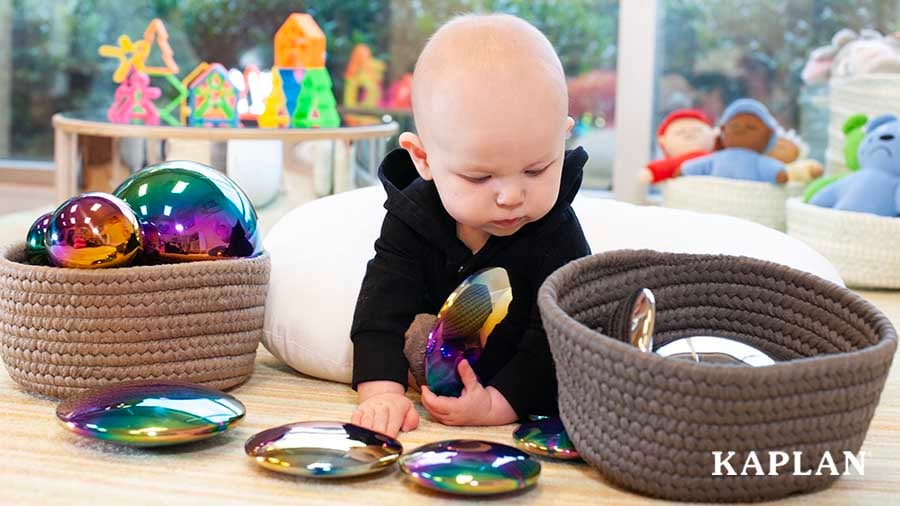 An infant is sitting on the floor, holding round iridescent stones while looking at a brown, woven basket on the floor. 