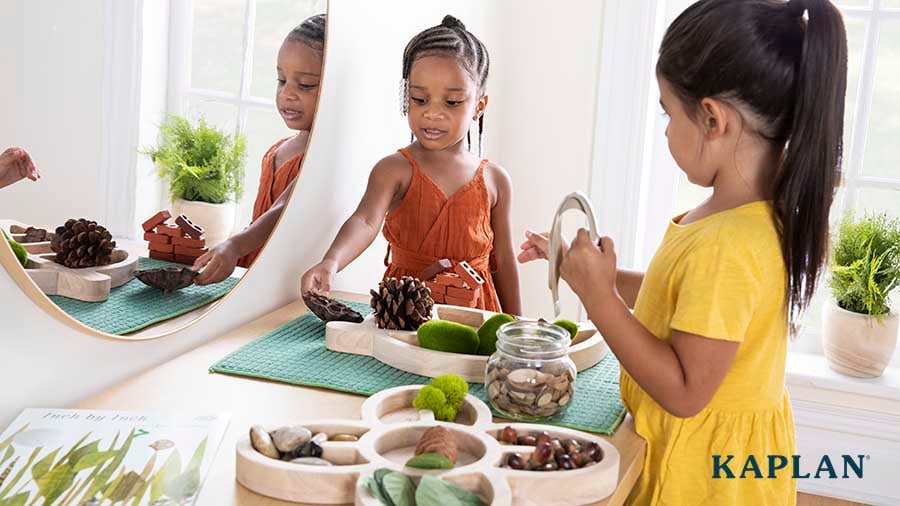 Two preschool children look down at a Wooden Loose Parts Tray containing various natural material such as a pinecone and moss. 