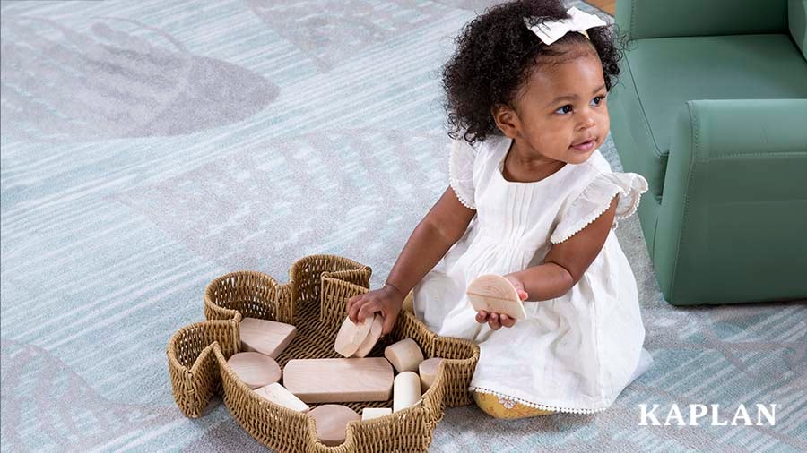A toddler girl in a white dress holds wooden blocks in both hands, while sitting beside a turtle-shaped Washable Wicker Basket that contains wooden blocks. 