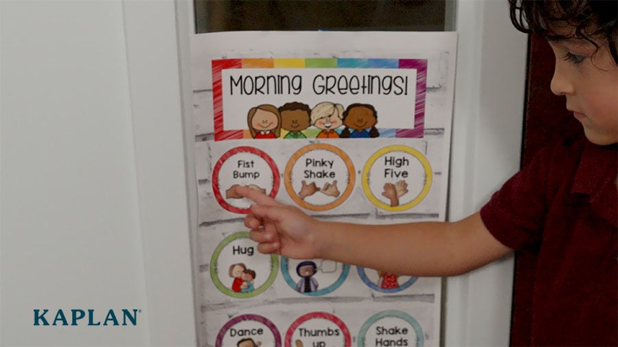 A young boy points to the "fist bump" image on a morning greeting chart located beside a preschool classroom door. 