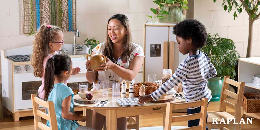 An early childhood teacher sits at a wooden table, she is holding a teapot in her hands. Three young children are gathered at the table. Two are looking at the teacher and the teapot, another child is placing a wooden egg in a pan which is placed on the table. 