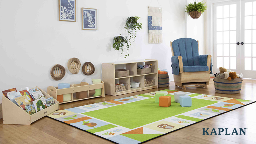 An image of an early childhood toddler classroom that is set up with a large area rug, bookshelf, wooden storage shelving, a wooden rocking chair with a blue cushion, a wicker basket with stuffed animals, and wall tapestries and planters on the walls. 