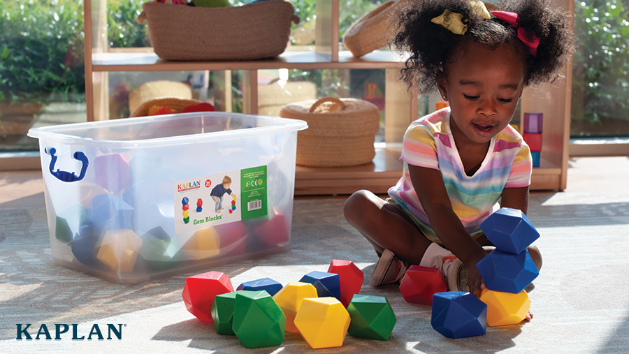 A toddler plays with gem shaped blocks in primary colors next to a reusable, plastic, storage container.
