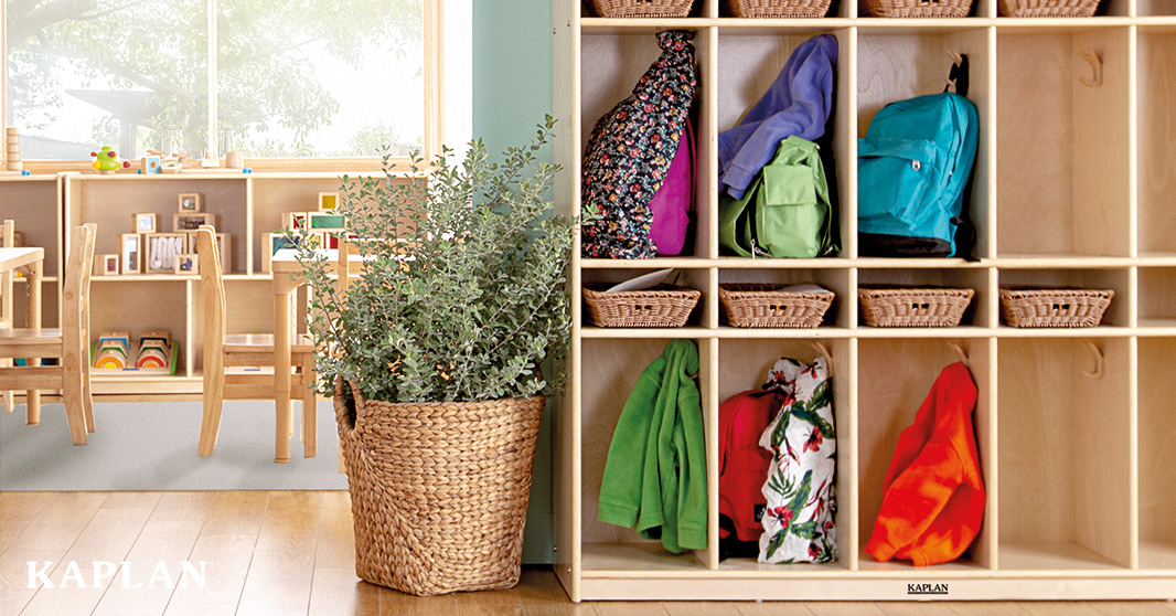 A Carolina Birch locker storage unit filled with children's backpacks and jackets and storage baskets. A large plant beside the storage unit bridges the doorway into the the main classroom space filled with birch tables, chairs, and storage units.