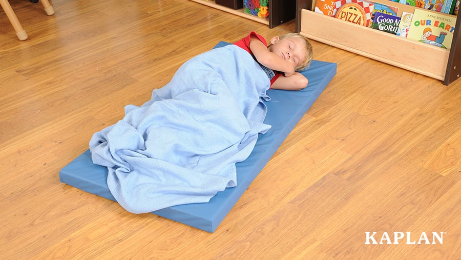 A preschool-aged boy takes a nap on a blue mat on a classroom floor