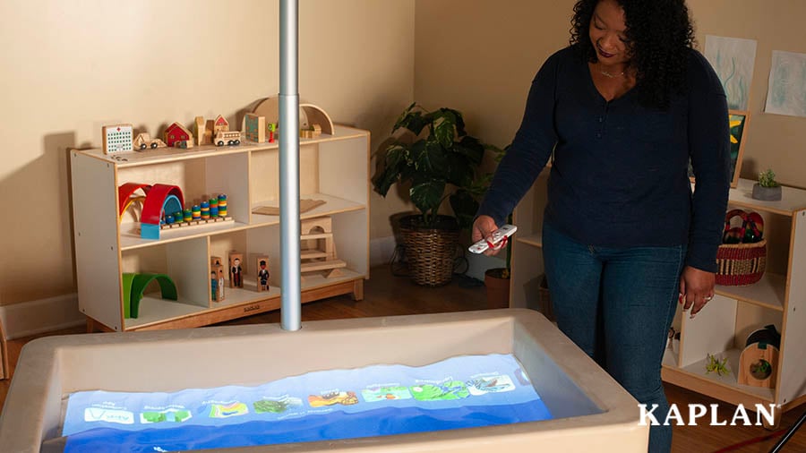 A female teacher, wearing a black sweater, holding a silver remote in her right hand, stands beside of a sand table lit up with a blue screen projecting activity options for sand play. 