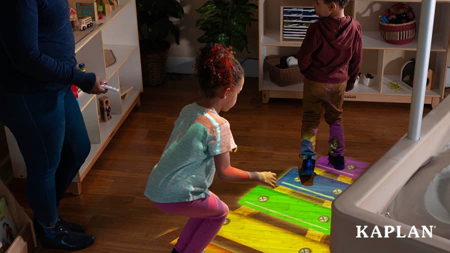 A young boy and a young girl take turns hopping across a colorful xylophone projected onto the wood floor, while a female teacher controls the projector with a remote control. 