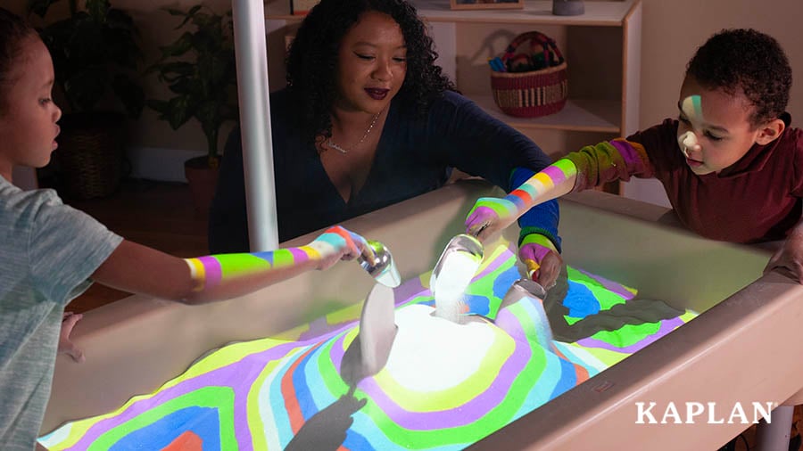 Two young children and a female teacher lean over a sand table lit up with colorful circle projections. Each of them are holding a metal shovel, while scooping sand out of the table and pouring it back inside the table. 