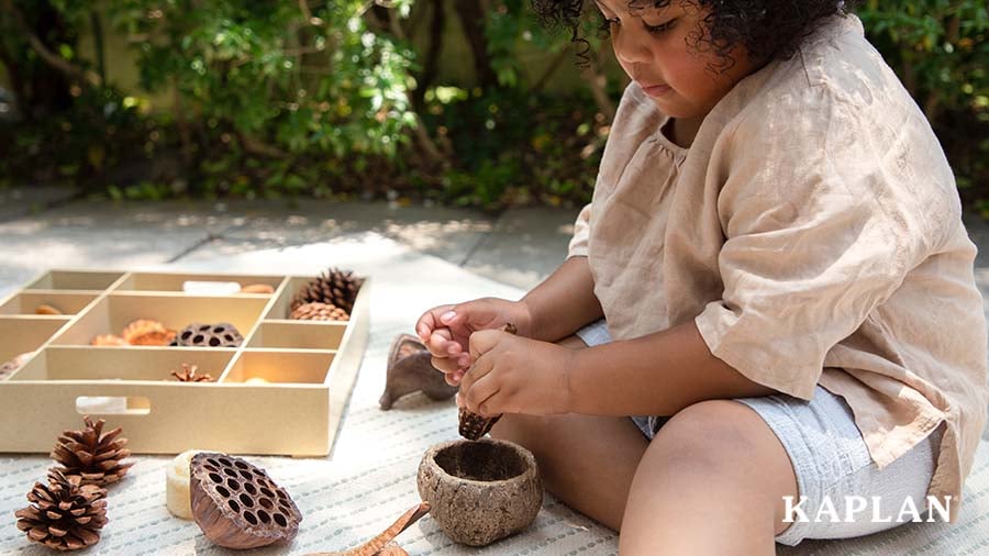 A young girl sits on a blanket outside, a natural loose parts kit is in front of her. She is holding a shell with her left hand, while looking down at a wooden bowl in front of her on the blanket. 