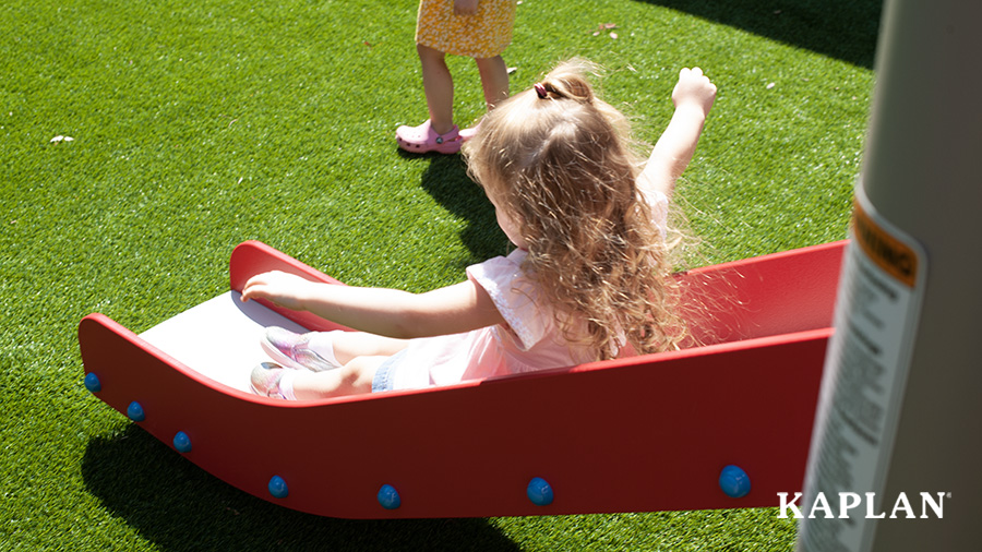 A toddler slides down a red slide over a synthetic green grass surface.