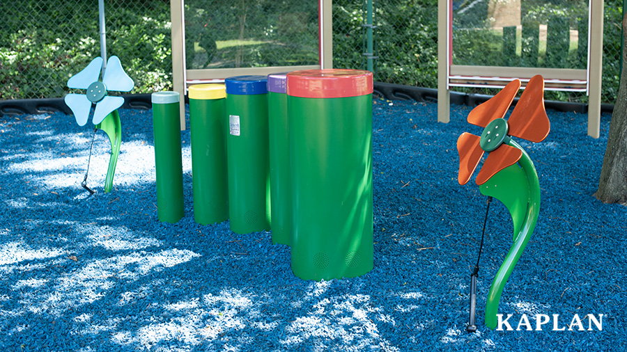 Several metal musical instruments set up on a rubber mulch playground surface, in the shade.
