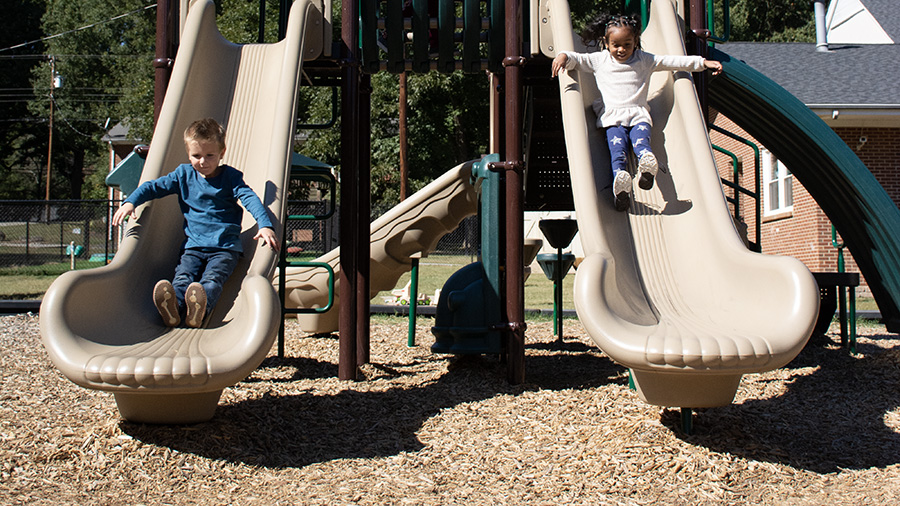 Two preschool children slide down double slides on playground equipment that is set on a wooden mulch surface.