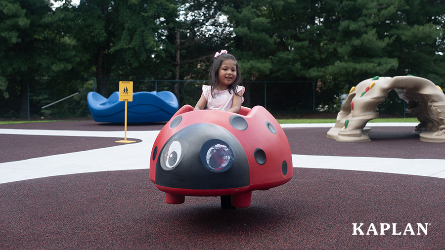 A young girl in a pink shirt sits in a red ladybug bouncer at the playground.