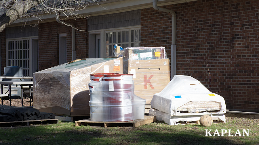 Playground equipment sits outside, on pallets, wrapped in cardboard and plastic wrap.