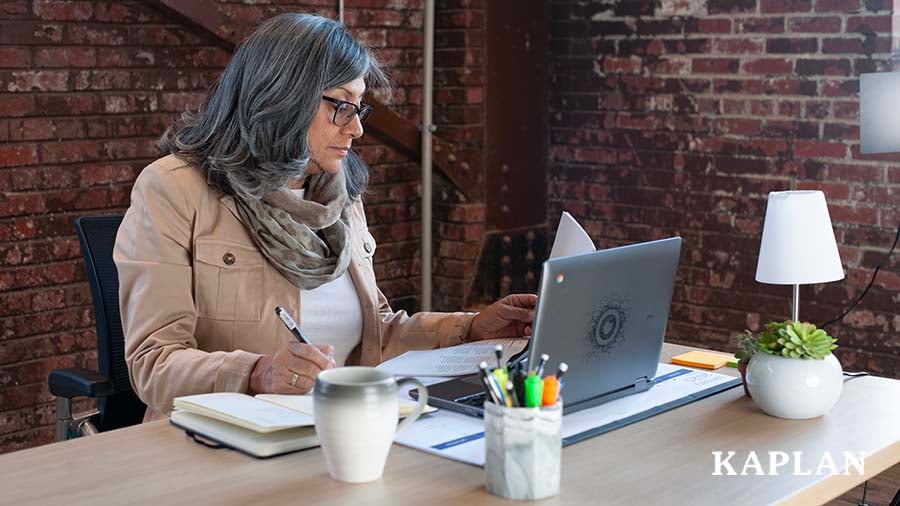 A female early childhood professional sits at a desk in front of her computer, while holding an ink pen in her right hand, looking at a piece of paper that is in her left hand.