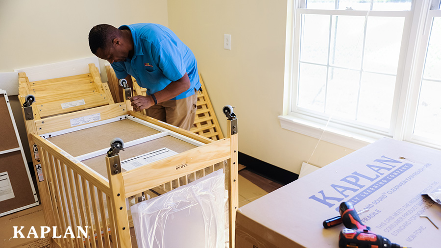 A deliveryman in a blue shirt assembles an upside-down, wheeled, crib, using various tools, in a classroom filled with delivery boxes. 