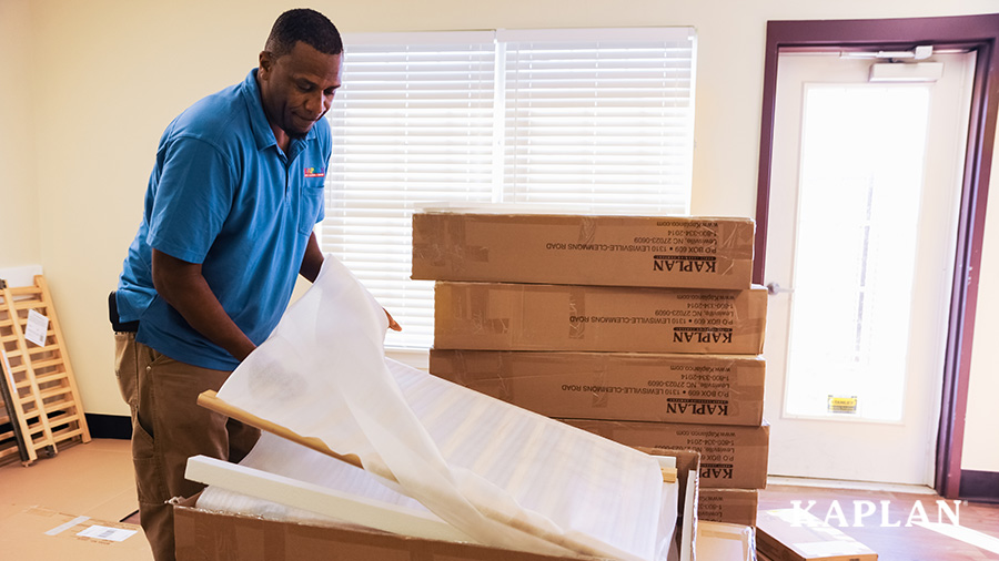 A deliveryman in a blue shirt carefully unwraps and removes the foam protecting the sides of a wooden crib packed inside a Kaplan delivery box.