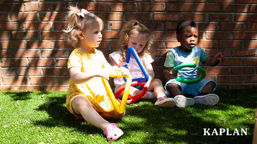 Three young children sit on a patch of grass, beside a red brick wall, in the shade, each are holding plastic connecting shape toys.