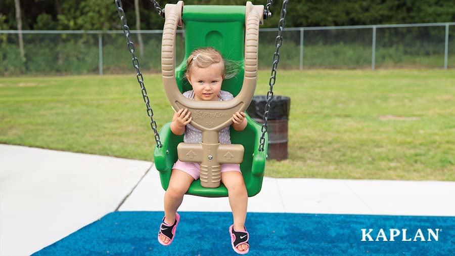 A young child wearing a grey t-shirt, pink shorts, and black sandals sits in a green inclusive swing.