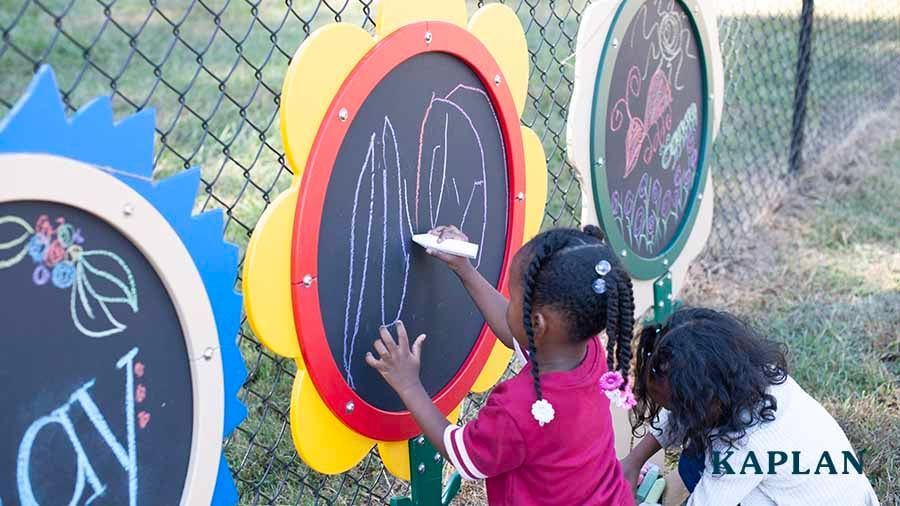 A flower-shaped chalkboard hangs on a chainlink fence, two young girls stand in front of it, holding large chalk pencils, drawing lines on the chalkboard.