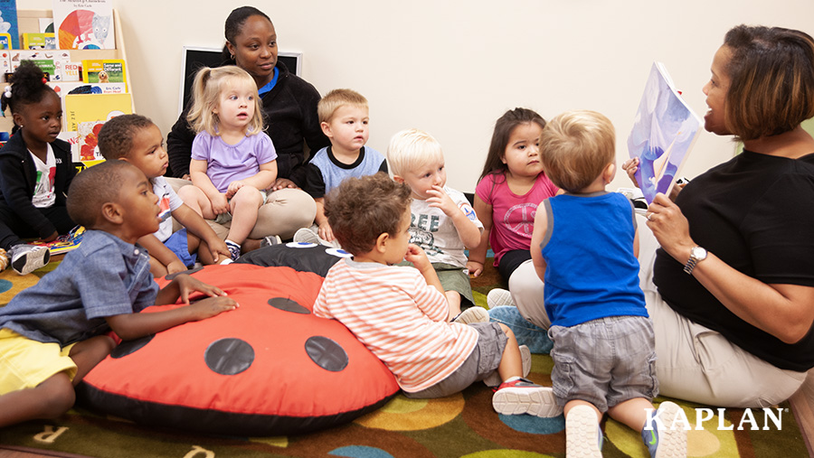 A female teacher sits on the floor of her classroom, holding up a book while reading to a group of children who gather around, looking at the teacher.