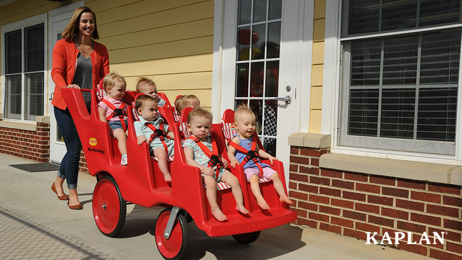 A female teacher pushes a group of infants in a red, six-passenger, buggy, on a sunny playground.