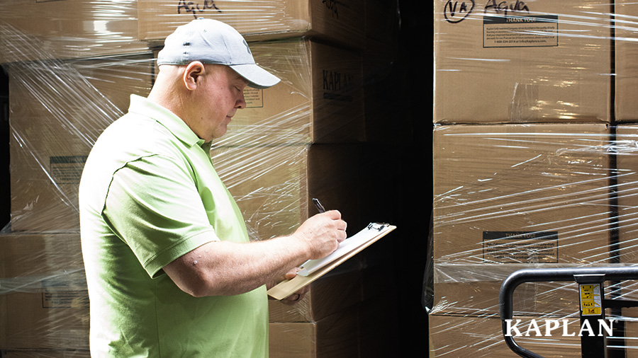 A delivery driver, standing next to a stack off boxes, checks off inventory on a clipboard.