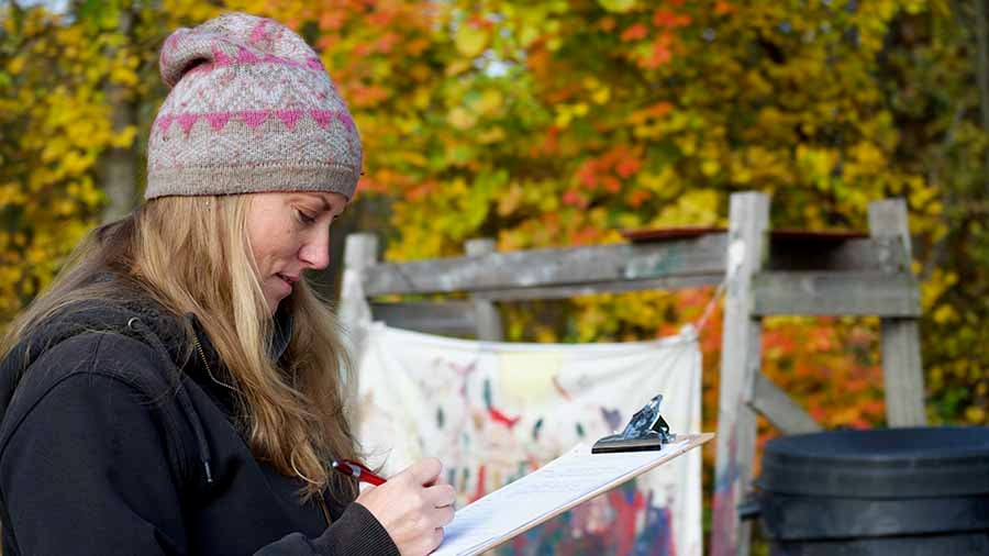 A female teacher wearing a pink and grey beanie is standing outdoors, holding a clipboard with a piece of paper attached to it. She is writing on the paper with a red pen. 