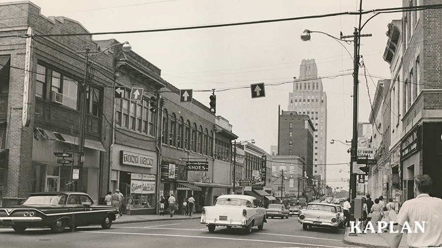 An image of a city street in Greensboro, NC in 1951, cars are pictured on the road, and the Tiny Town toy store sign is seen in the background. 