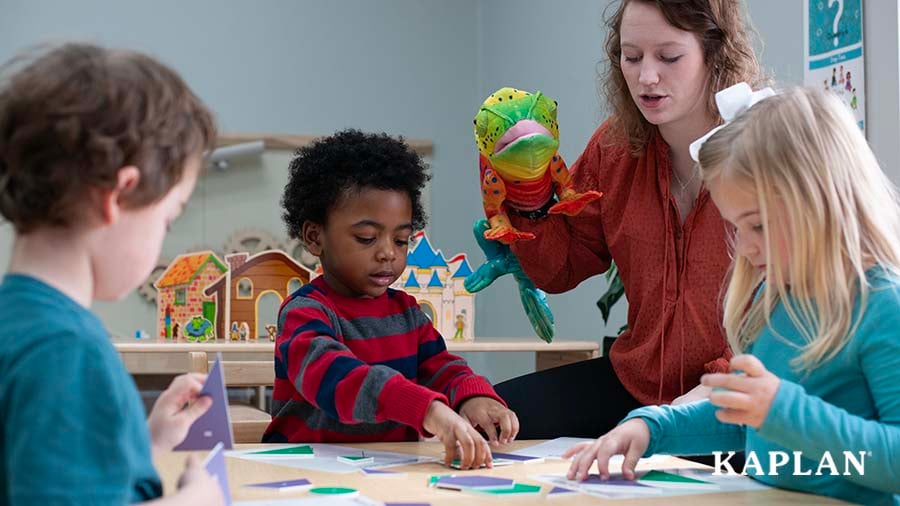 A teacher sits at a table, a chameleon puppet is on her right hand. Three children are siting around the table, using their hands to pick up wooden shapes in various sizes.