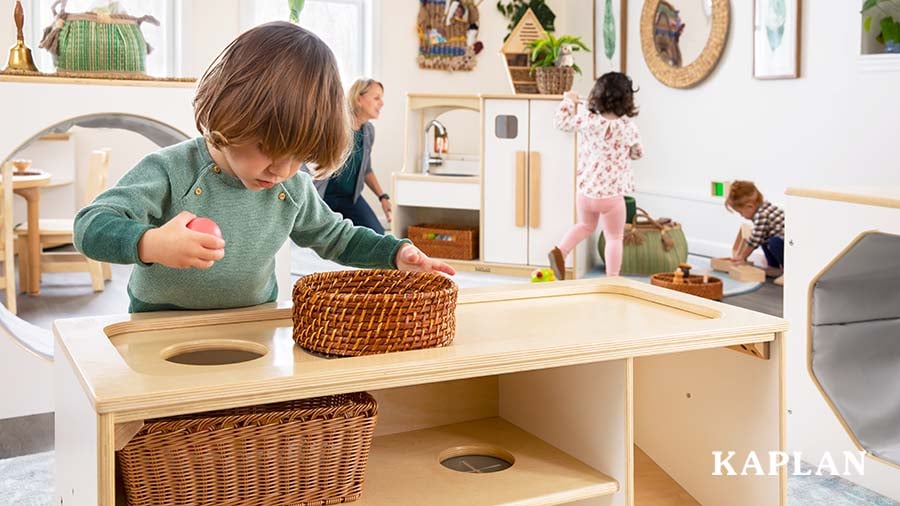 A young child in a green shirt stands at the Sense of Place for Wee Ones Activity Stand, holding a pink ball in one hand while looking down through a hole constructed on the top of the shelving unit. 