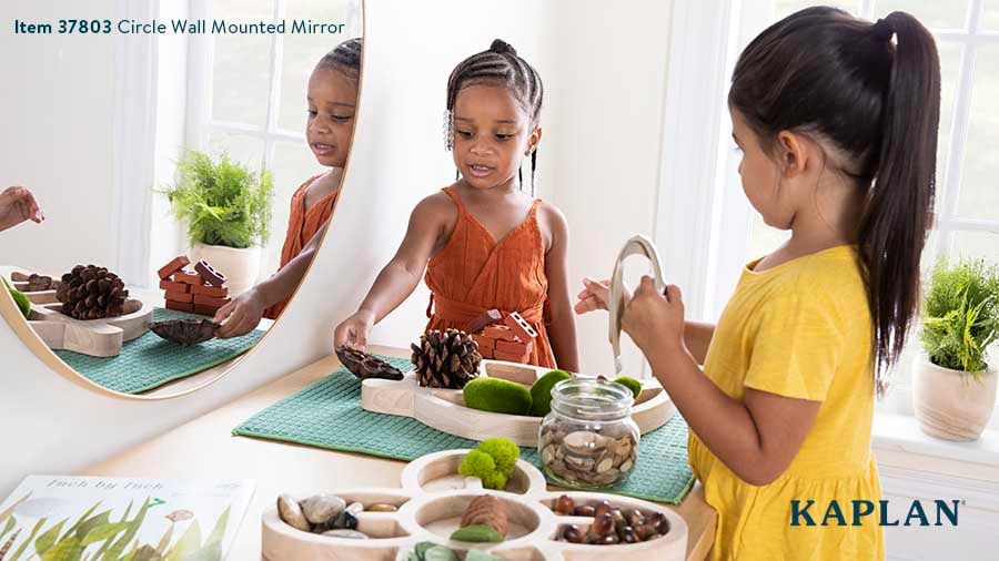 Two young children stand in front of a Circle Wall Mounted Mirror, while looking at nature-inspired materials on a table positioned below the mirror. 