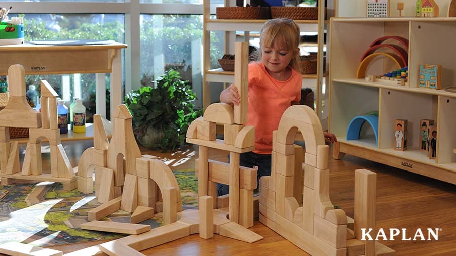 A young child in an orange shirt sits on the floor in a pre-K classroom while placing blocks one on top of the other to create different shapes and buildings. 