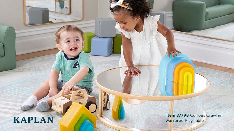 Two toddlers play on the floor of an early childhood classroom, the Curious Crawler Mirrored Play Table is in front of them. The toddler girl stands at the table while playing with Rainbow Arch blocks. 