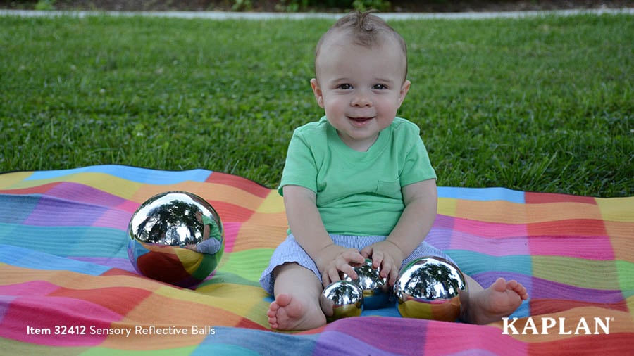 A young infant wearing a green shirt sits on a multi-colored blanket outdoors, while holding a Sensory Reflective Ball by Kaplan. 