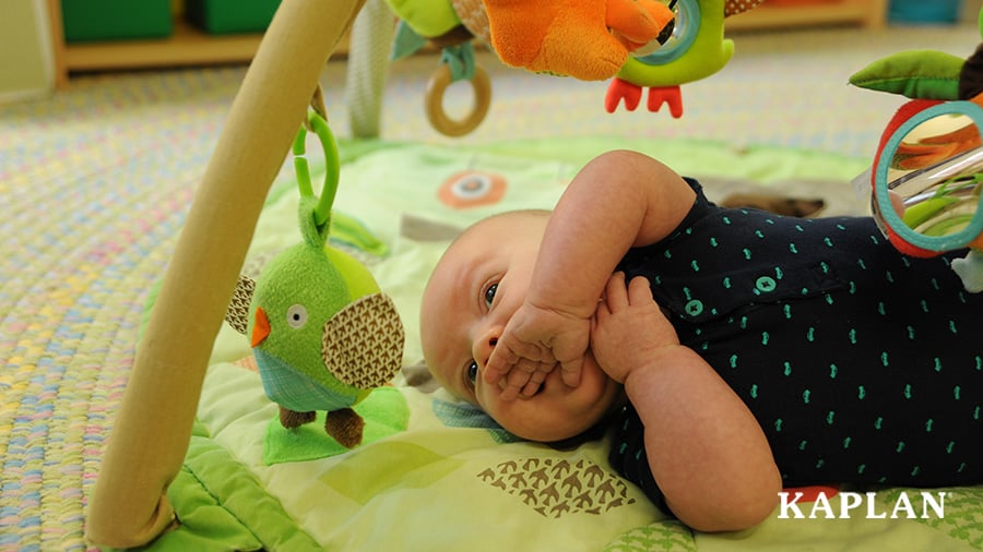 An infant lays on a green play mat with plush birds hanging from the arches, that is placed on top of a woven pastel rug in an infant classroom. 